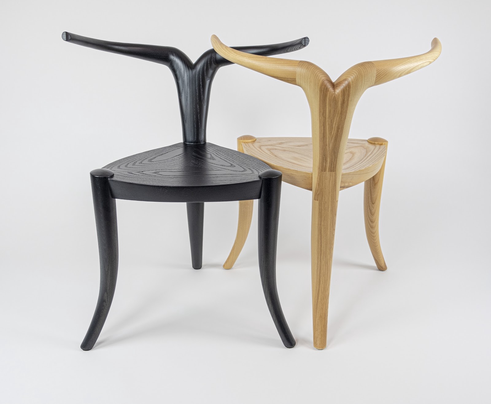 Two 3-legged wood chairs, one natural, one black. For a back, each has a single pole that splits into two antler-like arms.