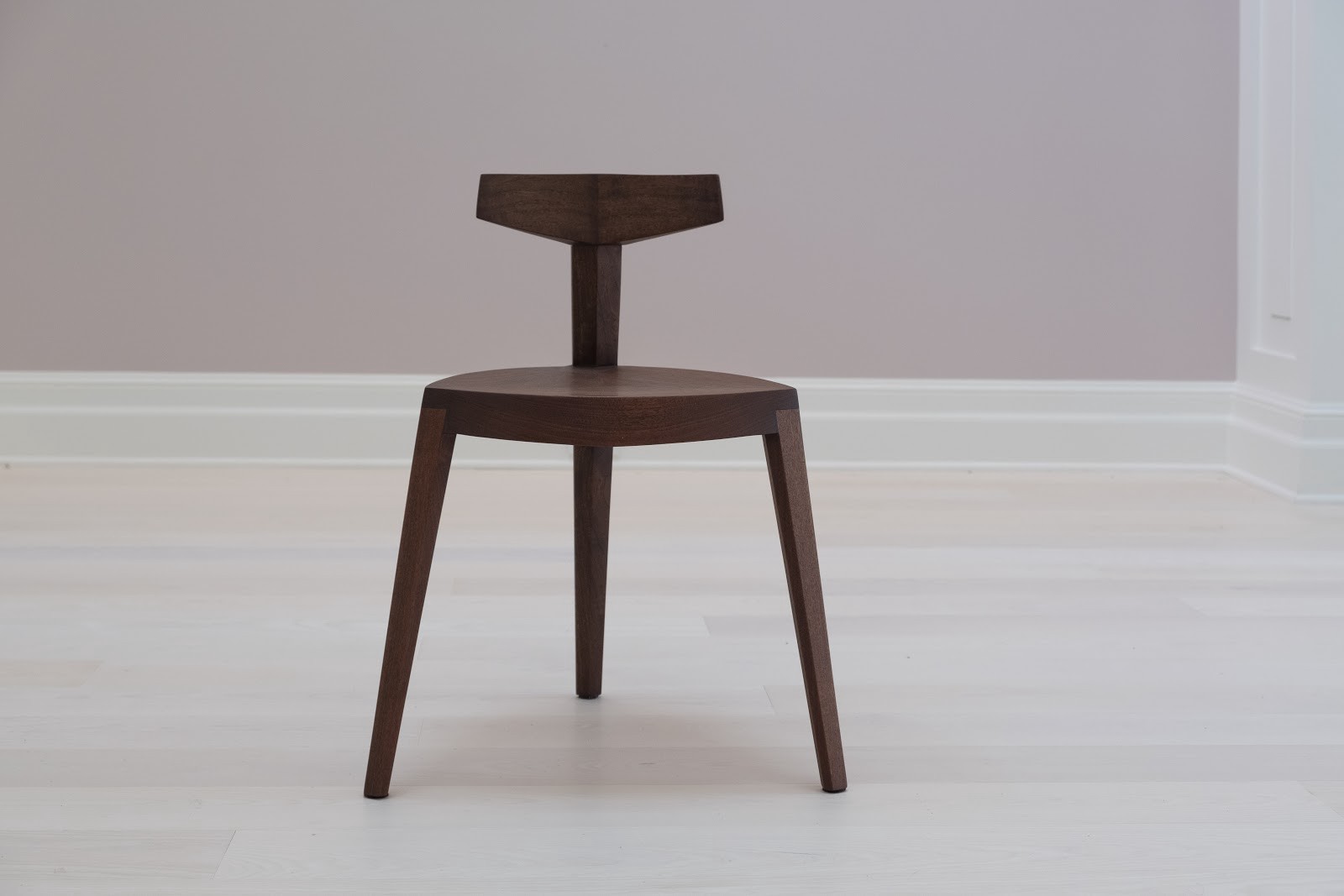 Brown wooden chair with 3 angular legs and a short back made from a vertical rectangular pole with a crossbar at the top.