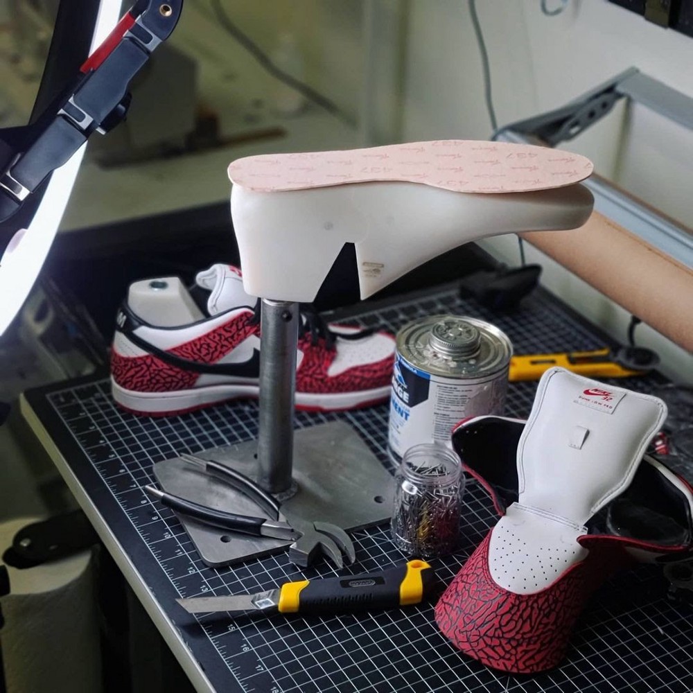 On a workbench a short pole holds a solid form shaped like a right sneaker upside down. Lying on it is part of a footbed.