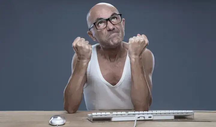 Man in an undershirt at a desk balls fists and looks upward in anger as if cursing the skies