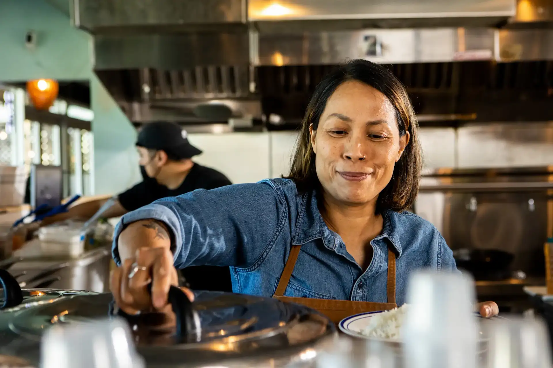 Smiling person at work in a professional kitchen holds a bowl with rice and lifts a pot lid as they plate a meal.