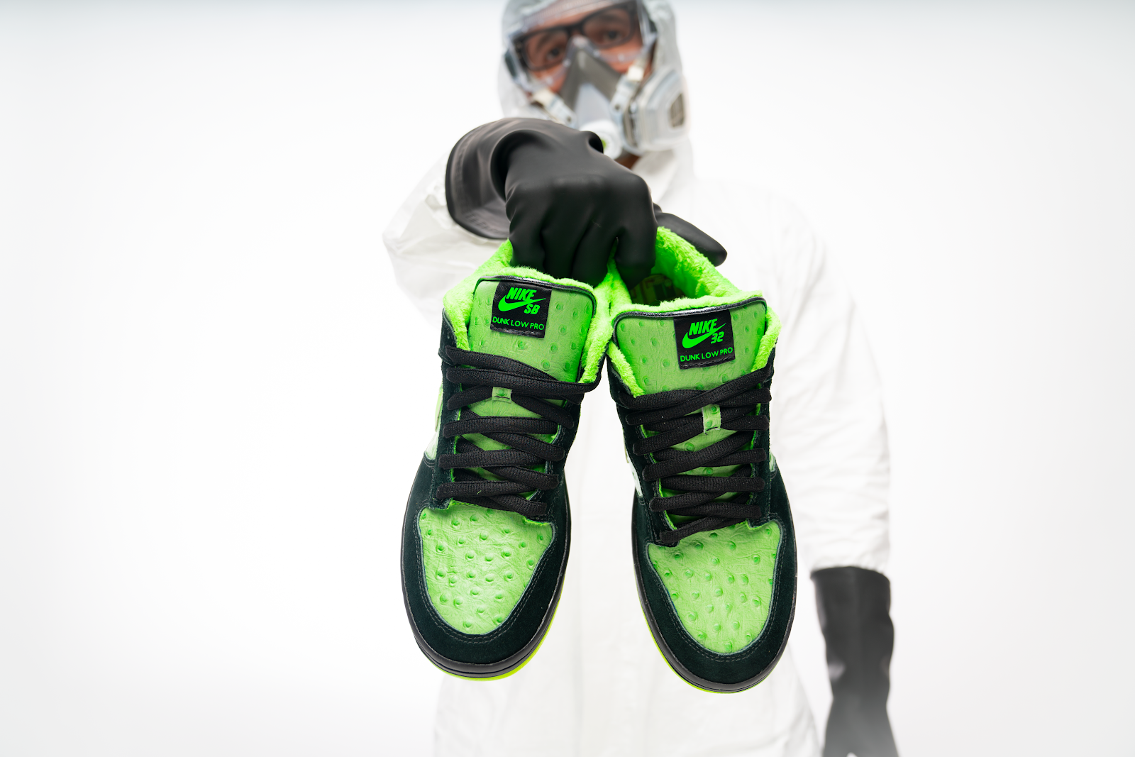 Barrios wears a hazard suit, face mask, and gloves while holding a pair of black and neon-green low-top Nikes.